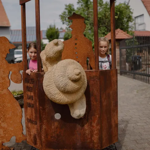 Zwei Kinder stehen in einer rostigen Schuldenfigur auf einem Gehweg. Im Hintergrund sind ein paar Häuser und Bäume zu sehen.