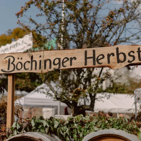 Ein Holzschild mit der Aufschrift "Böchinger Herbst" steht im Vordergrund. Im Hintergrund sind Bäume und eventuelle Marktstände zu sehen.