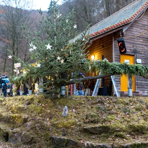 Ein rustikales Holzhaus umgeben von einer malerischen Landschaft. Vor dem Haus steht ein geschmückter Weihnachtsbaum mit Sternen und eine fröhliche Menschenmenge versammelt sich.