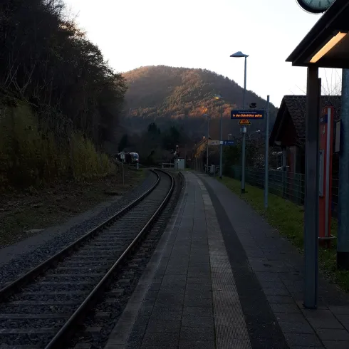 Ein ruhiger Bahnhof in der Natur mit Gleisen und einem Blick auf einen Hügel im Hintergrund. Die Uhr und die Signaltafeln sind sichtbar.