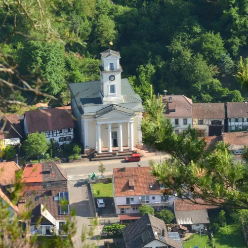 Eine malerische Stadtansicht mit einer Kirche im Zentrum und umliegenden Wohnhäusern. Grünes Land und Bäume umgeben die Szene.