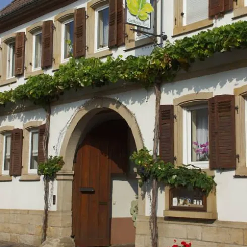 Eine charmante Fassade eines Hauses mit Holztüren und Fensterläden. Über dem Eingang hängt ein Schild, und Weinreben umschlingen die Wände.