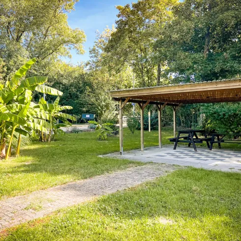 Ein ruhiger Garten mit einer überdachten Sitzgelegenheit und viel Grün. In der Nähe stehen große Pflanzen und Bäume.