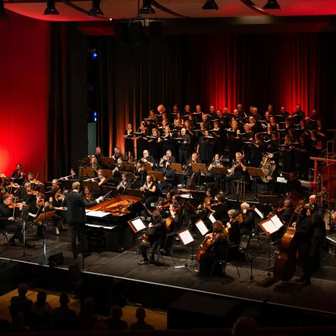 Ein Orchester und ein Chor treten auf einer Bühne mit roter Beleuchtung auf. Die Musiker konzentrieren sich auf ihre Instrumente und die Performance.