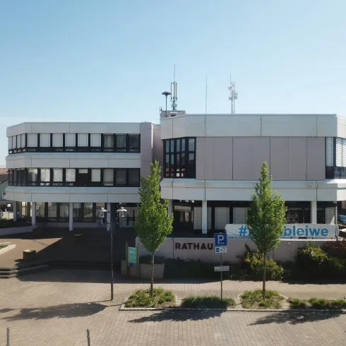 Ein modernes Gebäude mit klaren Linien und großen Fenstern. Es gibt Bäume und eine gepflasterte Fläche vor dem Eingang.