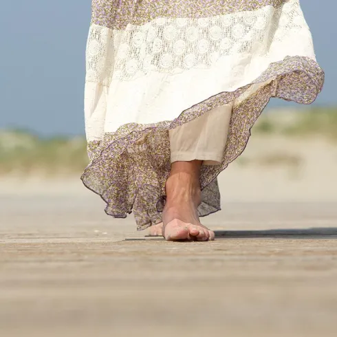 Eine Person mit einem langen, fließenden Kleid geht barfuß über eine Holzplanke. Im Hintergrund sind sanfte Dünen und ein blauer Himmel sichtbar.