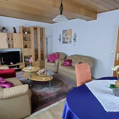 Ein gemütliches Wohnzimmer mit beigen Sofas und einem runden Tisch. Der Raum ist hell und einladend, mit dekorativen Elementen und einem Fernseher.