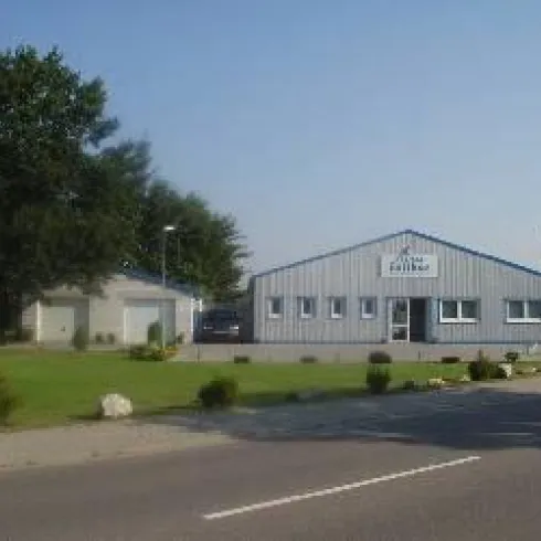 Ein modernes Gebäude mit blauer Dachfarbe und großen Fenstern steht an einer Straße. Daneben befindet sich ein kleineres Nebengebäude und ein gepflegter Rasen.