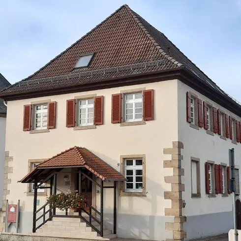 Ein charmantes, zweigeschossiges Gebäude mit einem schrägen Dach und roten Fensterläden. Der Eingangsbereich ist über eine kleine Treppe mit einem Vordach zugänglich.