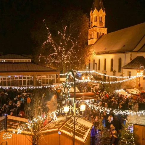 Ein festlicher Weihnachtsmarkt bei Nacht mit vielen Besuchern. Glänzende Lichter und eine Kirche im Hintergrund schaffen eine gemütliche Atmosphäre.