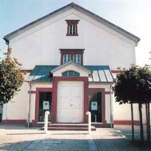 Ein modernes Gebäude mit einem weißen Eingang und einem symmetrischen Dach. Vor dem Haus stehen mehrere Bäume und der Weg ist gepflastert.