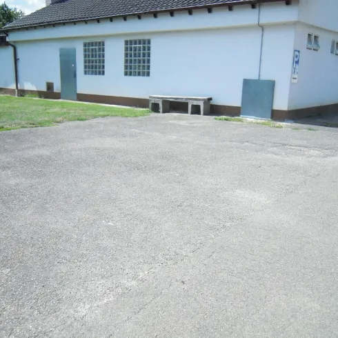 Ein leerer, asphalierter Platz neben einem weiß gestrichenen Gebäude. Im Hintergrund sind grüne Wiesen und ein blauer Himmel zu sehen.