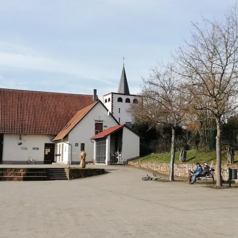 Ein ruhiger Platz mit einem modernen Gebäude und einer Kirche im Hintergrund. Im Vordergrund sind Bäume und Sitzbänke zu sehen.