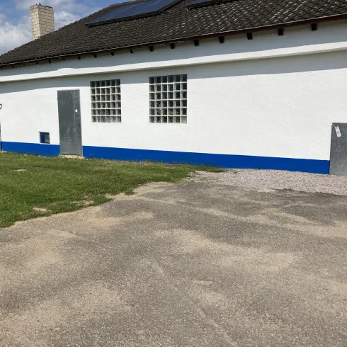 Ein einfaches Gebäude mit einer weißen Wand und blauen Akzenten. Der Boden ist befestigt und es gibt einen grünen Rasen davor.