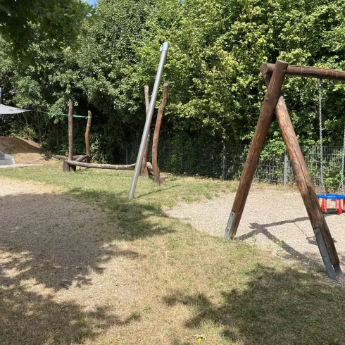 Ein Spielplatz mit Schaukeln und Holzstrukturen. Umgeben von Bäumen und einer grünen Wiese.