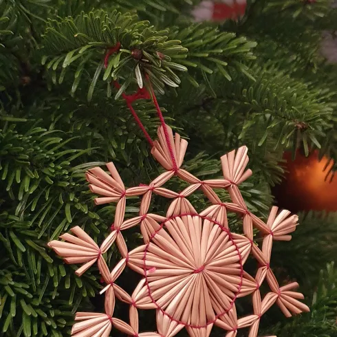 Ein kunstvoller Weihnachtsschmuck in Form einer Schneeflocke hängt an einem grünen Tannenzweig. Die Verzierung ist aus geflochtenem Stroh und zeigt feine Details.