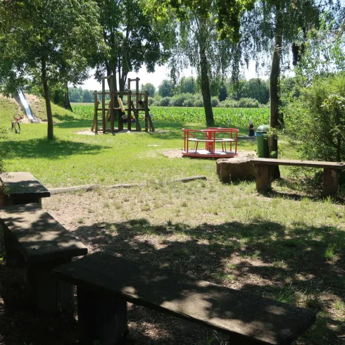 Ein schöner Spielplatz mit einer Rutsche und einem Klettergerüst, umgeben von Bäumen. Im Hintergrund sind grüne Felder zu sehen.