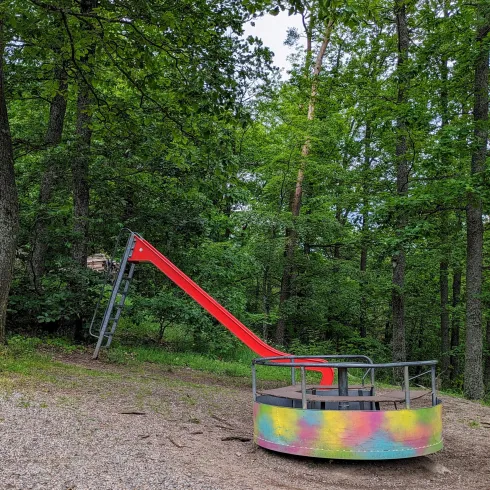 Ein bunter Spielplatz im Wald mit einer rotfarbenen Rutsche und einer Drehplattform. Die Umgebung ist von hohen Bäumen umgeben.