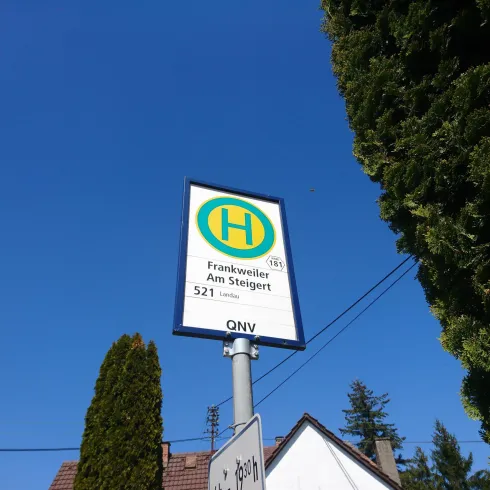 Ein Haltestellenschild mit der Aufschrift „Friedrichstraße Am Staupert“ vor einem klaren blauen Himmel. Umgeben von Bäumen und einer Wohngegend.