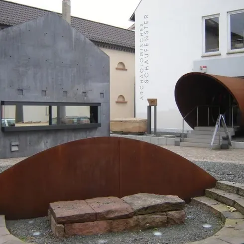Ein moderner Platz mit verschiedenen architektonischen Elementen. Im Vordergrund befindet sich eine runde, metallische Skulptur und im Hintergrund sieht man zwei Gebäude mit unterschiedlichen Baumaterialien.