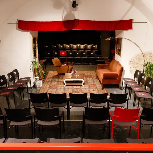 Ein gemütlicher Veranstaltungsraum mit Stühlen in Reihen und einem kleinen Wohnzimmerbereich. Im Hintergrund befindet sich eine Bühne mit rotem Vorhang und dekorativen Elementen.