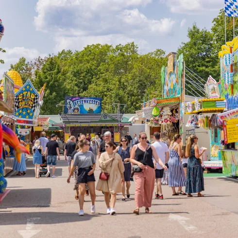 Ein belebter Jahrmarkt mit bunten Ständen und vielen Besuchern. Im Hintergrund sind grüne Bäume und ein blauer Himmel zu sehen.