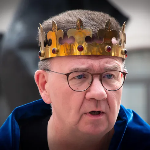 Ein Mann trägt eine goldene Krone mit roten Edelsteinen. Er hat einen ernsten Gesichtsausdruck und trägt einen blauen Umhang.