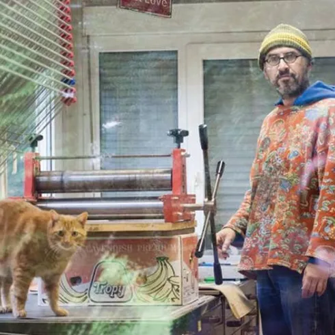 Ein Mann steht in einem kreativen Atelier neben einer orangefarbenen Katze. Im Hintergrund sieht man eine Druckmaschine und einen hellen Raum.