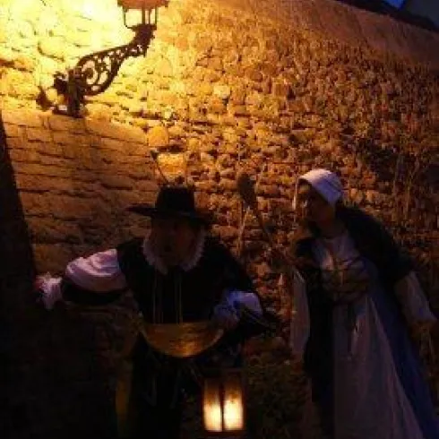 Zwei Menschen in historischen Kostümen stehen an einer alten Steinmauer. Eine Laterne beleuchtet die Szene in der Dämmerung.