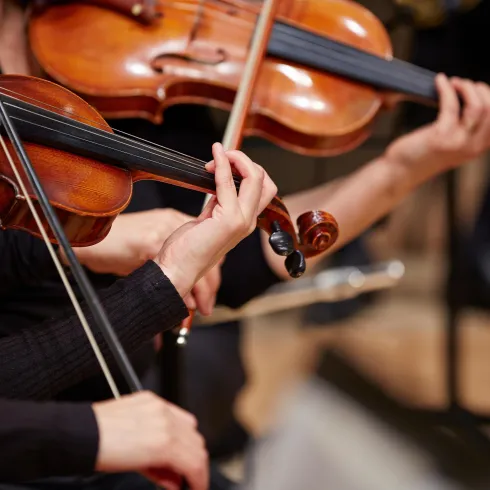 Nahaufnahme von Händen, die Geigen spielen. Die Stimmung ist musikalisch und konzentriert.