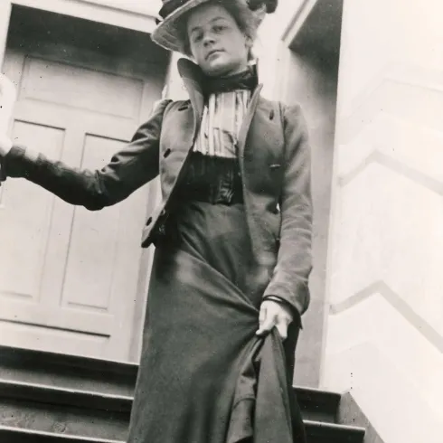 Eine elegant gekleidete Frau steht auf einer Treppe. Sie trägt einen auffälligen Hut und hat einen selbstbewussten Ausdruck.