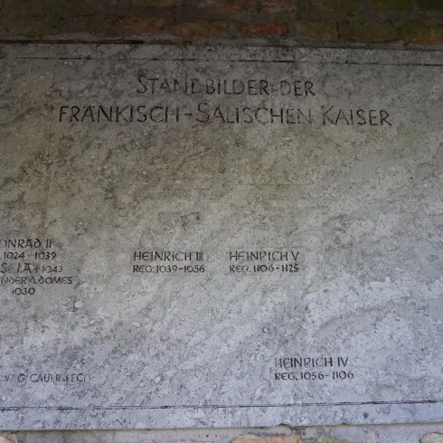 Eine Gedenktafel mit den Namen der fränkisch-salischen Kaiser. Die Tafel ist an einer Wand befestigt und aus grauem Stein.