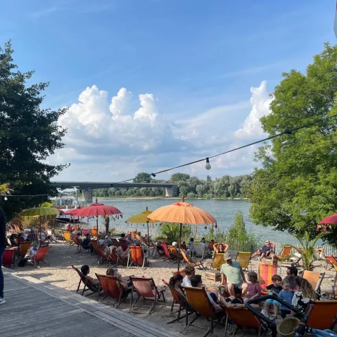 Ein schöner Außenbereich am Fluss mit gemütlichen Liegestühlen und bunten Sonnenschirmen. Viele Menschen genießen die Atmosphäre und das Wetter.