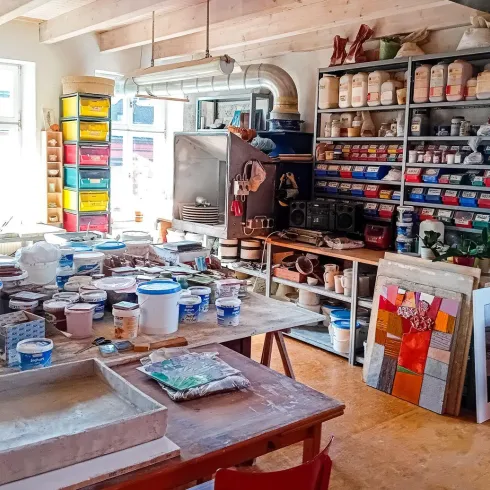 Ein kreatives Atelier mit Tischen voller Malutensilien und Farbtöpfen. Regale mit verschiedenen Kunstmaterialien und Farbpaletten füllen den Raum.