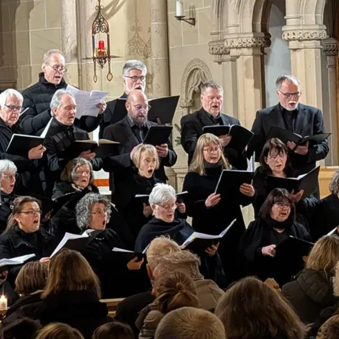 Ein Chor steht in einer Kirche und singt. Die Mitglieder tragen schwarze Kleidung und haben Notenblätter in der Hand.