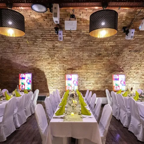 Ein eleganter Veranstaltungsraum mit langen Tischen, die mit weißen Tischdecken und grünen Servietten dekoriert sind. Die warmen Lichtakzente schaffen eine einladende Atmosphäre.