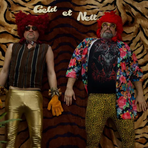 Zwei Männer in auffälligen, bunten Outfits liegen auf einem gestreiften Hintergrund. Sie tragen rote Perücken und Accessoires, die einen humorvollen Eindruck vermitteln.