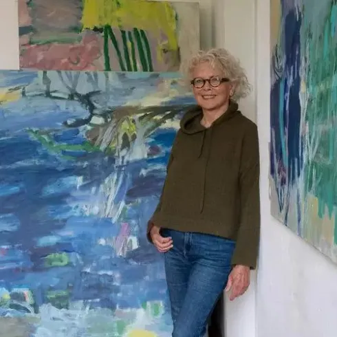Eine Frau steht in einem Kunststudio und lächelt. Im Hintergrund sind mehrere farbenfrohe Gemälde zu sehen.