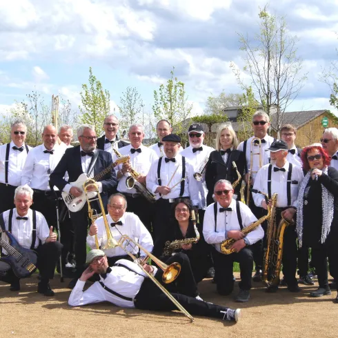 Eine große Gruppe von Musikern in formeller Kleidung, die verschiedene Instrumente halten. Sie stehen in einem Garten mit bunten Blumen und einem schönen Himmel im Hintergrund.