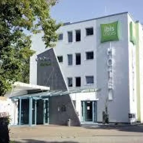 Ein modernes Hotelgebäude mit einer klaren Fassade und einem Eingangsbereich. Umgeben von Bäumen und unter blauem Himmel.