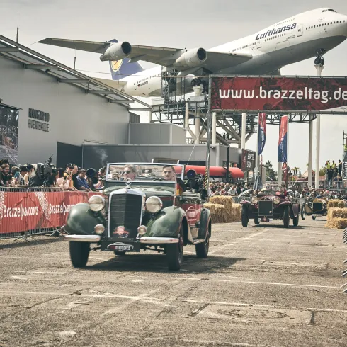 Eine Oldtimer-Rallye findet auf einem Flughafen statt. Im Hintergrund fliegt ein Flugzeug über die Rennstrecke.