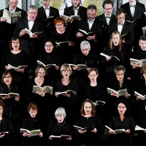 Ein großer Chor mit vielen Sängern in schwarzen Kleidungen steht zusammen auf einer Bühne. Alle halten Notenblätter und singen gemeinsam.
