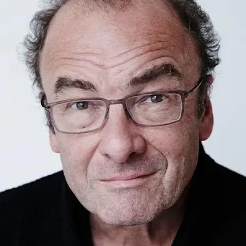 Ein Mann mit Brille und leichtem Lächeln schaut in die Kamera. Er hat graues Haar und trägt ein schwarzes Oberteil.