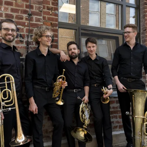 Eine Gruppe von fünf Musikern steht vor einer Backsteinwand. Sie tragen schwarze Kleidung und halten verschiedene Blasinstrumente.