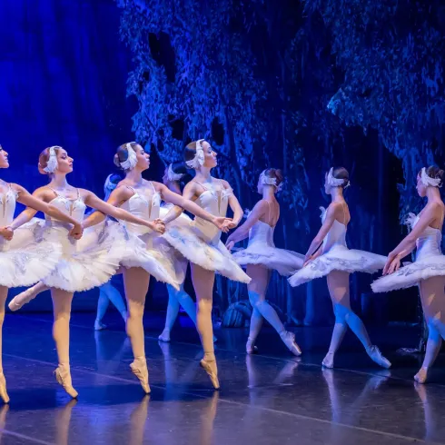 Eine Gruppe von Ballerinen in weißen Tutus tanzt auf einer Bühne. Der Hintergrund ist blau beleuchtet, was eine elegante Atmosphäre schafft.