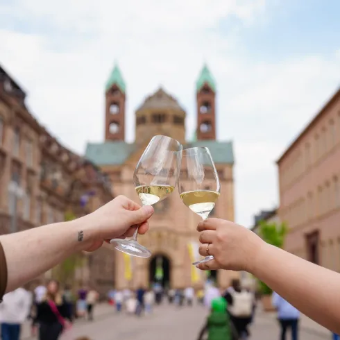 Zwei Personen stoßen mit Gläsern Sekt an, während im Hintergrund eine historische Kirche zu sehen ist. Der Platz ist belebt und es herrscht eine festliche Stimmung.