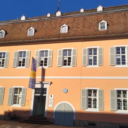 Ein pastelgelbes Gebäude mit weißen Fensterläden und einem Satteldach. Das Haus hat eine elegante Fassade und steht unter einem klaren blauen Himmel.