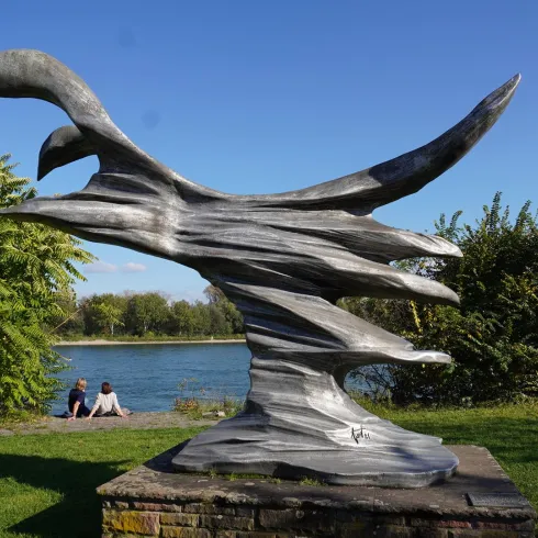 Eine moderne Skulptur, die spiralförmig gestaltet ist und an Bewegung erinnert. Im Hintergrund sind Besucher am Wasser zu sehen.