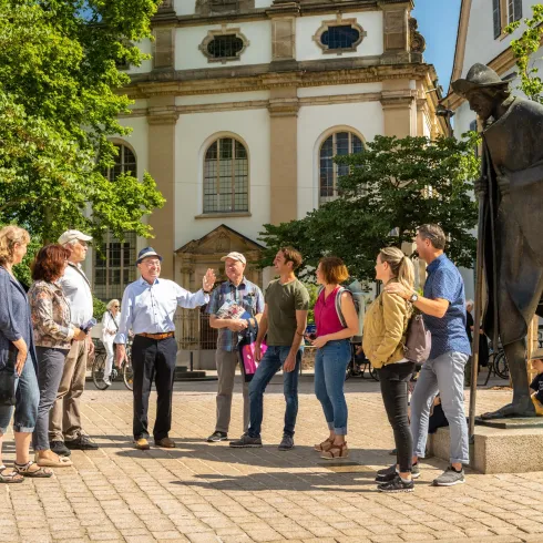 Eine Gruppe von Touristen hört einem Reiseführer zu, während sie auf einem Platz stehen. Im Hintergrund ist eine Statue und historische Gebäude zu sehen.