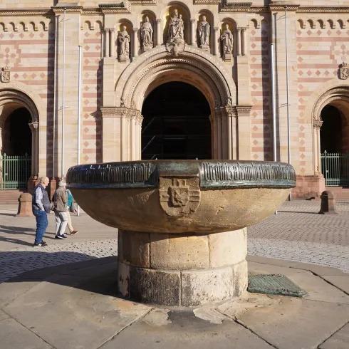 Ein historischer Brunnen steht im Vordergrund, umgeben von Menschen, die vorbeigehen. Im Hintergrund ist eine beeindruckende Gebäude­fassade mit Bögen und kunstvollen Skulpturen zu sehen.